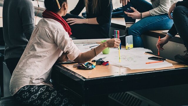 Frau lehnt aufeinen Tisch mit Papieren und wirkt als wäre sie in eine Diskussion vertieft, mit Menschen die nicht in gänzezu sehen sind.