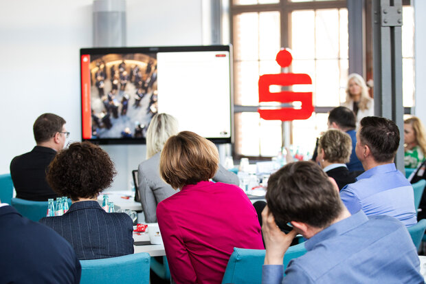 Menschen sitzen in einem Sparkassenseminar. Im Hintergrund ist ein Sparkassenlogo