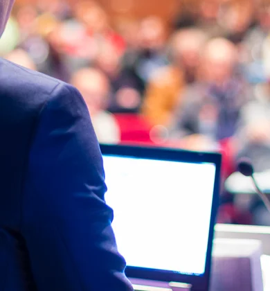 Ein Sprecher steht mit dem Rücken zur Kamera vor einem Publikum und hält vermutlich eine Rede, vor ihm steht ein Laptop und ein Mikrofon auf einem Sprecherpult