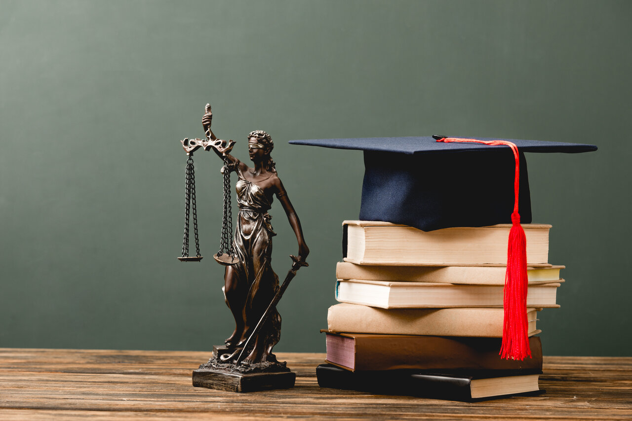 Bild einer Justizia Statue und einem Stapel Bücher mit einer amerikanischen Graduationskappe vor einem grünen Hintergrund auf einem Holztisch.